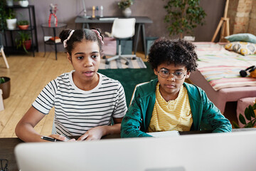 Two African school children sitting at table in front of computer monitor and having online meeting with teacher during onine studying