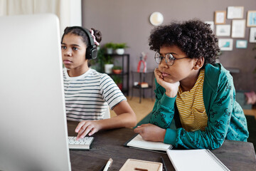 African boy in eyeglasses looking at computer monitor while girl playing video game on computer during their leisure time at home