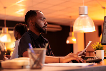 Business man analyzing paperwork report and taking notes to create research presentation in office with big windows. Working on project data with documents and computer during sunset.