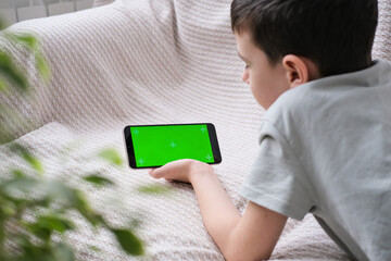 The boy looks at the screen of the phone with a chrome key indoors. An unrecognizable child uses a smartphone with a green screen to watch a video.