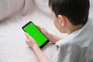 The boy looks at the screen of the phone with a chrome key indoors. An unrecognizable child uses a...