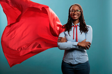 Brave and happy looking young adult superhero woman wearing red hero cape while smiling at camera. Justice defender with superpowers and mighty posture standing with arms crossed on blue background. - obrazy, fototapety, plakaty