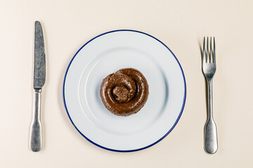 Plastic excrement lies on a plate between a fork and a knife close-up. The concept of an inept cook, fake news, shitty restaurant, bad food, taste of shit