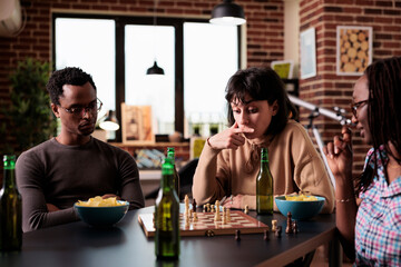 Pensive woman sitting at table while thinking about next chess move. Young smart looking people playing strategy boardgames together while sitting at home in living room.