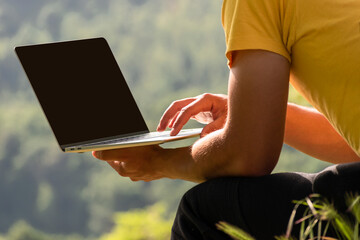 Young man using laptop computer, notebook outside in nature against background of mountain forest. Online remote working and learning. Copy space for design or text on laptop black screen