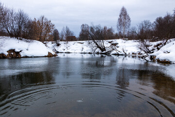 forest river with fast flow and small waves flows between snowy shores with trees and bushes late winter and early spring