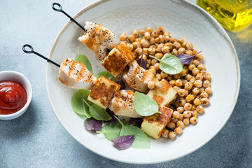 Grilled chicken meat and halloumi cheese on skewers served with roasted chickpeas in a beige plate,...