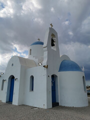 The Church of St. Nicholas the Wonderworker is white with a blue door and a bell against the backdrop of the Mediterranean Sea and the dramatic sky.