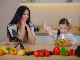 Happy mother and daughter enjoy prepare freshly salad together in kitchen. Healthy food at home. Healthy Lifestyle and Eating Concept. little influencer filming blog about healthy eating