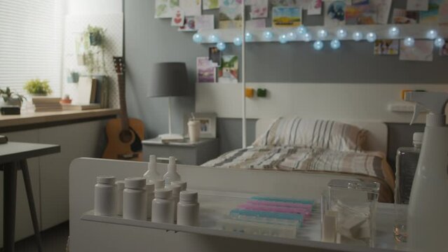 View of plain pill jars and disposable supplies on table in decorated hospital ward of child or teenager with garland, guitar, many pictures, drawings and postcards on wall daytime