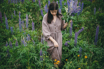 Cottagecore aesthetics. Stylish woman in rustic dress holding lupine bouquet in meadow. Young...