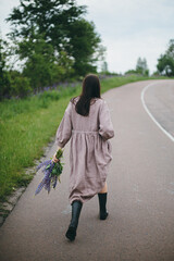 Stylish woman in rustic dress walking with lupine bouquet in summer countryside. Cottagecore aesthetics. Young female in linen dress holding wildflowers after rain on rural road, slow life