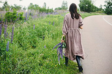 Stylish woman in rustic dress walking with lupine bouquet in summer countryside. Cottagecore...