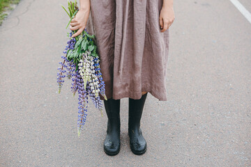 Cottagecore aesthetics. Close up of woman in rustic dress and gumboots holding lupine bouquet in summer countryside. Young female with wildflowers after rain on rural road. Moody image