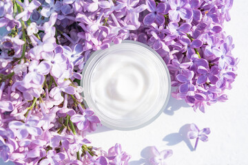 Opened white jar of herbal light cream for women on white table. Beautiful branch of purple lilac. Care for clean and soft face, hands, legs and body skin. Fresh flowers. Empty place for text.