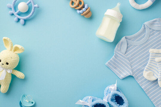 Baby accessories concept. Top view photo of infant clothes blue shirt socks knitted bunny toy bottle teether rattle shoes and pacifier on isolated pastel blue background with empty space in the middle