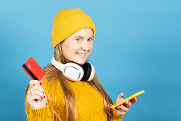 Portrait of cute teenager girl holding credit card and phone isolated on blue background. Young woman online shopping