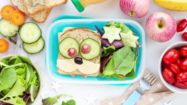 Back to school concept. Lunch box preparation for kids with a cheese and salami sandwich, fresh salad leaves, cucumbers, carrots and tomatoes in stop motion animation