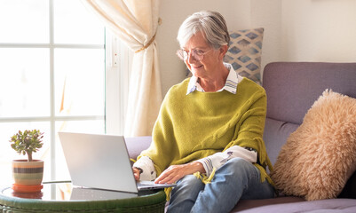 Smiling elderly woman working on laptop at home sitting on sofa in living room. Modern senior lady...