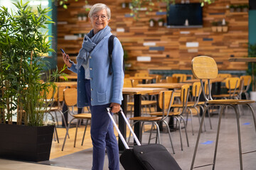 Smiling caucasian traveler senior woman with backpack and hand luggage walking in airport waiting...