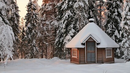 A small wooden barbecue hut. Behind the hut, there are big snowy spruces. It is a beautiful winter evening.