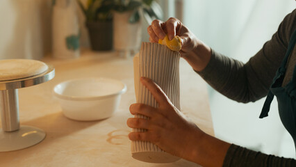 Detailed Frame of Caucasian Woman's Hands Holding a Clay Vase While She Goes Over It with a Wet Sponge. Wooden Surface for Working.