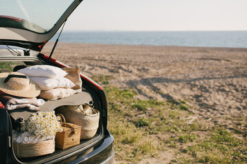 Open trunk of car with picnic items: straw hat, basket with food and flowers with summer beach on...