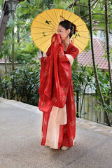 Fototapeta premium Young attractive Asian woman wearing tadeonal Chinese red hanfu long skirt dress costume scarf hairpin earing decorated umbrella outdoor green garden patio