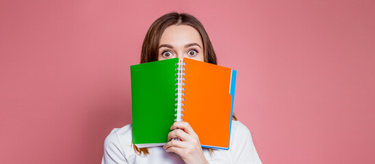 Shocked young girl student hides her face behind a notebook and surprised holds an exercise book in...