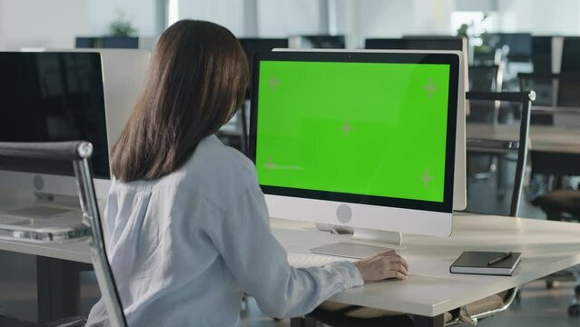 Creative Young Woman Sitting at His Desk Using Desktop Computer with Mock-up Green Screen. Evening in the Stylish Office Studio. Beautiful Diverse Female Manager Surfing Internet in Green Screen
