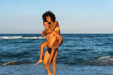 Happy young interracial couple in piggybak on beach – man giving piggybak his woman and laughing...