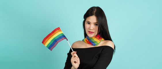 LGBTQ woman holding pride flag standing against a blue green background. Asian LGBTQ woman with rainbow scarf on neck. look smart bright and energetic cheerful. LGBTQ diversity pride concept.