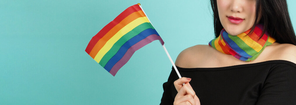 LGBTQ woman holding pride flag standing against a blue green background. Asian LGBTQ woman with rainbow scarf on neck. look smart bright and energetic cheerful. LGBTQ diversity pride concept.