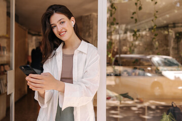 Fototapeta premium Pretty young caucasian woman looking at camera, holding phone in her hands standing in doorway. Brunette wears casual clothes in spring. Concept of use