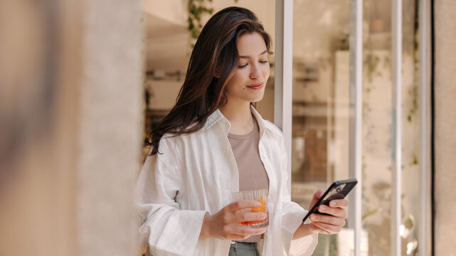 Pretty young caucasian woman is browsing email standing in doorway with glass of juice. Brunette wears top, shirt and pants. Cell phone usage concept
