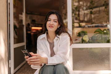 Pretty young caucasian woman with modern phone looking at camera sitting outdoors during day. Brunette wears casual clothes. Technology concept