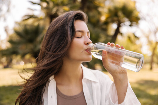 Pleasant young caucasian girl drinks refreshing water from bottle outdoors. Brunette with closed eyes wears white shirt. Concept of enjoying moment