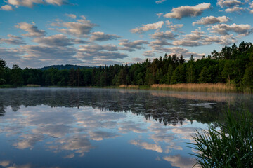 Fototapeta na wymiar Sunrise on the pond in the mountains. Rudawy Janowickie. Clouds in the sky. Clouds are reflected in the water. Around the forest. Mountains in the background. Fog over the water.