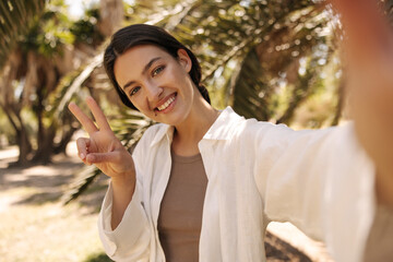 Fototapeta premium Pretty young caucasian girl smiling looking at camera, showing peace sign standing outdoors. Brunette with collected hair wears white shirt. People sincere emotions lifestyle concept.