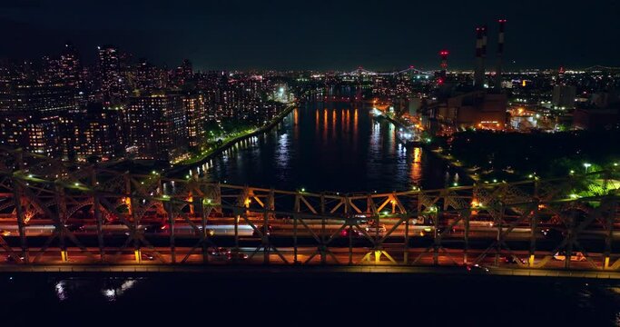 Multiple cars crossing the bridge at night. City lights reflecting in the river water. New York cityscape at backdrop.