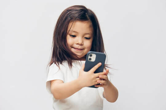 a cute little preschool girl stands on a white background in a white T-shirt and takes pictures of herself on her phone, smiling happily at the camera. The theme of children's happiness