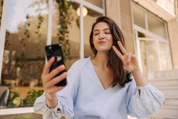 Cool young caucasian woman showing peace gesture in smartphone screen photographing herself. Brunette wears blue summer sundress. Vacation, happiness and pleasure concept