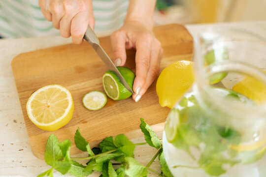 Hands of a woman preparing homemade mint, lemon, lime lemonade and a glass jug of lemonade. Cutting a lime on a wooden board for homemade lemonade.