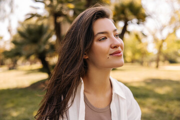 Fototapeta premium Close-up tender image of young caucasian woman enjoying sunny warm day in park. Brunette in shirt looks away. Lifestyle concept