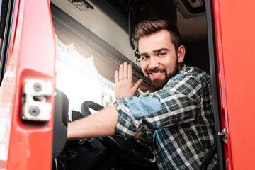 Smiling male truck driver sitting inside truck and waving with his hand