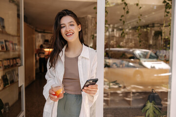 Positive young caucasian girl holding phone and glass with cocktail, looking at camera standing in doorway. Brunette wears casual summer clothes. Relaxation concept, technology