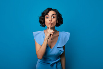 Close-up photo of a beautiful girl in a blue dress, who is looking in the camera while pressing her index finger to her lips and isolated on blue background