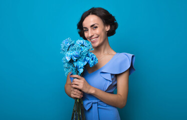 Young gorgeous woman in a bright blue dress is looking in the camera with a big smile, holding a bunch of blue flowers in her hands. International Women's Day