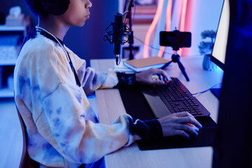 Cropped portrait of young teenage boy recording podcast at night in room with blue neon lighting,...