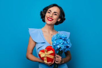 Women's day. St. Valentine's day. Close up portrait of elegant fashionable smiling woman in dress with bunch of flowers and gift box in hands isolated on blue background